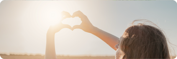 Sun, a source of vitamin D, in the sky with a woman looking up at the blue sky making a love heart shape with her hands around the sun.