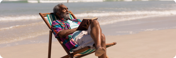 A dark skinned older man sitting in a deck chair reading a book on a beach to represent that people with darker skin need to spend longer in the sun to produce the same amount of vitamin D as someone with lighter skin.