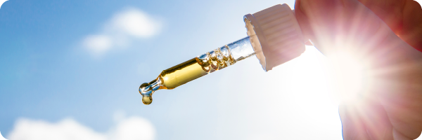A person's hand holding vitamin D liquid supplement up against a blue sky and sunny background.
