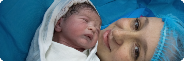 A mother in a hospital cap lying down holding a newborn baby wrapped in a white blanket to represent the fact that being born by caesarean section can affect the gut microbiome.
