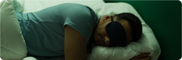 A man sleeping in bed in darkness wearing an eye mask. Sleeping in total darkness can improve sleep.