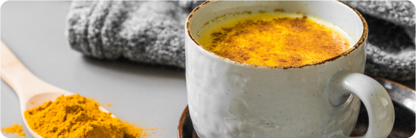 Turmeric milk my improve sleep. Image shows a cup of turmeric milk next to a grey blanket and a spoon overflowing with yellow turmeric powder. 