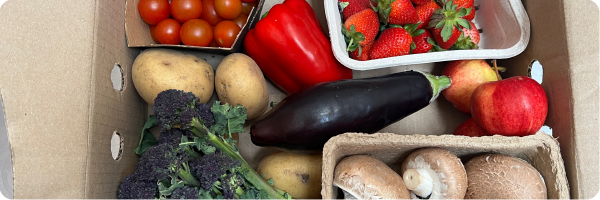 A cardboard box filled with potatoes, tomatoes, red peppers, strawberries, red apples, mushrooms, broccoli and aubergine. A vegetable box in one way to eat more vegetables.