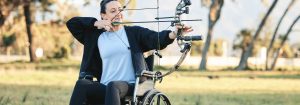 A Caucasian lady in a wheel chair getting ready to fire an arrow from a bow doing archery on a field outdoors on a bright day to represent Disability Pride Month.
