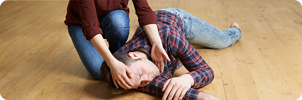 A Caucasian woman gently placing a Caucasian man's head in the recovery position with one ear to the ground. The man, wearing blue denim jeans and a red chequered shirt, appears unconscious and is lying on his side on a wooden floorboarded floor. 
