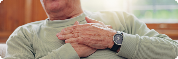 An older man in a pale green jumper has both of his hands on his chest, one on top of the other.