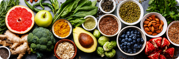 A table with brightly-coloured plant foods displayed: broccoli, blueberries, pomegranate, almonds, spinach, apple, turmeric, ginger, chia seeds, Brussel sprouts, oats, seeds, parsley, green chillis.