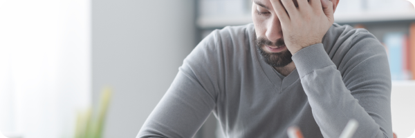 A bearded man in a long-sleeve, grey t-shirt looks tired. His left hand is propping up his head as he leans on a surface.