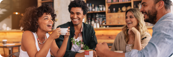 A group of young adults, including a black woman, black man, white woman and white man laughing and chatting in a cafe to represent finding support as you quit smoking. 