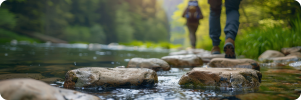 Two people's legs walking along stepping stones across a river with green trees in the background to represent that quitting smoking is about moving forward.