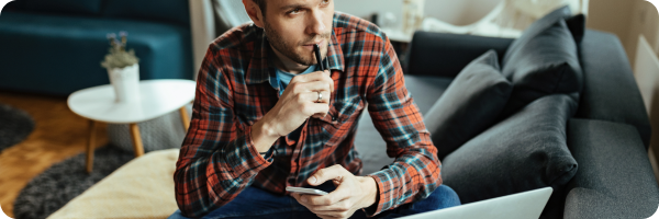A young man sat down in front of a laptop screen holding a pen to his lips and a notebook in the other hand looking thoughtful to represent common questions around nicotine replacement therapy. 