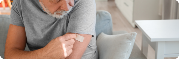 An old man in a grey short-sleeved t-shirt sitting down putting a nicotine patch on his arm. 